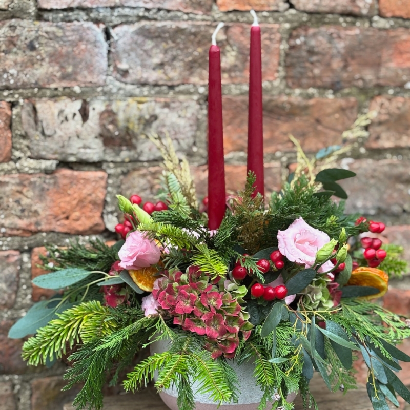 Christmas table arrangement Mulberry Blush with red candles pink roses hydrangea berries pine and festive foliage displayed in a ceramic bowl.