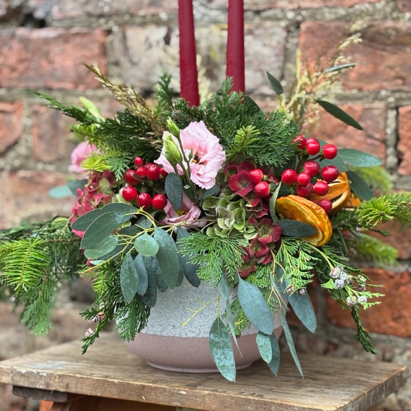 Christmas table arrangement Mulberry Blush with red candles pink roses hydrangea berries pine and festive foliage displayed in a ceramic bowl.