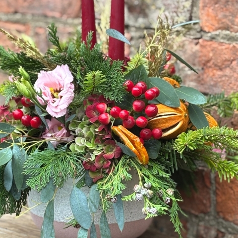 Christmas table arrangement Mulberry Blush with red candles pink roses hydrangea berries pine and festive foliage displayed in a ceramic bowl.