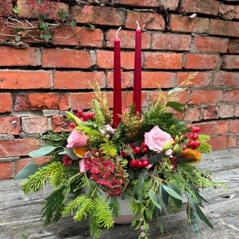 Christmas table arrangement Mulberry Blush with red candles pink roses hydrangea berries pine and festive foliage displayed in a ceramic bowl.