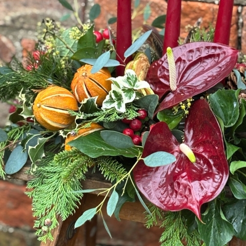 Noël Luxe Christmas table arrangement with red candles anthuriums dried oranges berries pine foliage and a pheasant feather displayed on a rustic table.