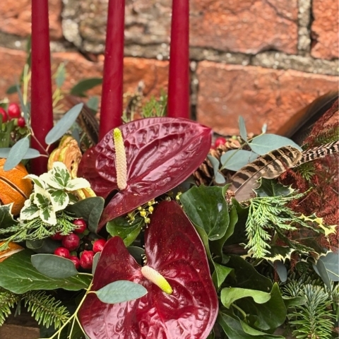 Noël Luxe Christmas table arrangement with red candles anthuriums dried oranges berries pine foliage and a pheasant feather displayed on a rustic table.