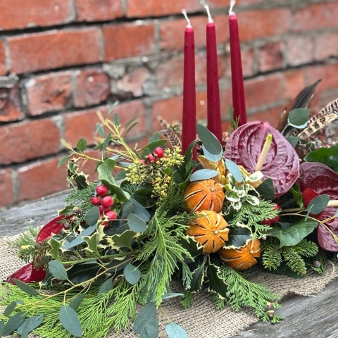 Noël Luxe Christmas table arrangement with red candles anthuriums dried oranges berries pine foliage and a pheasant feather displayed on a rustic table.