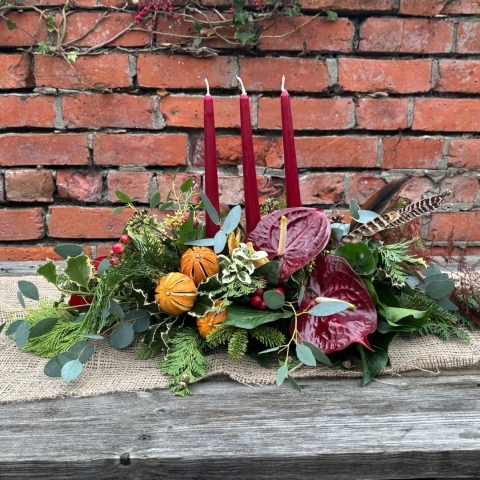 Noël Luxe Christmas table arrangement with red candles anthuriums dried oranges berries pine foliage and a pheasant feather displayed on a rustic table.